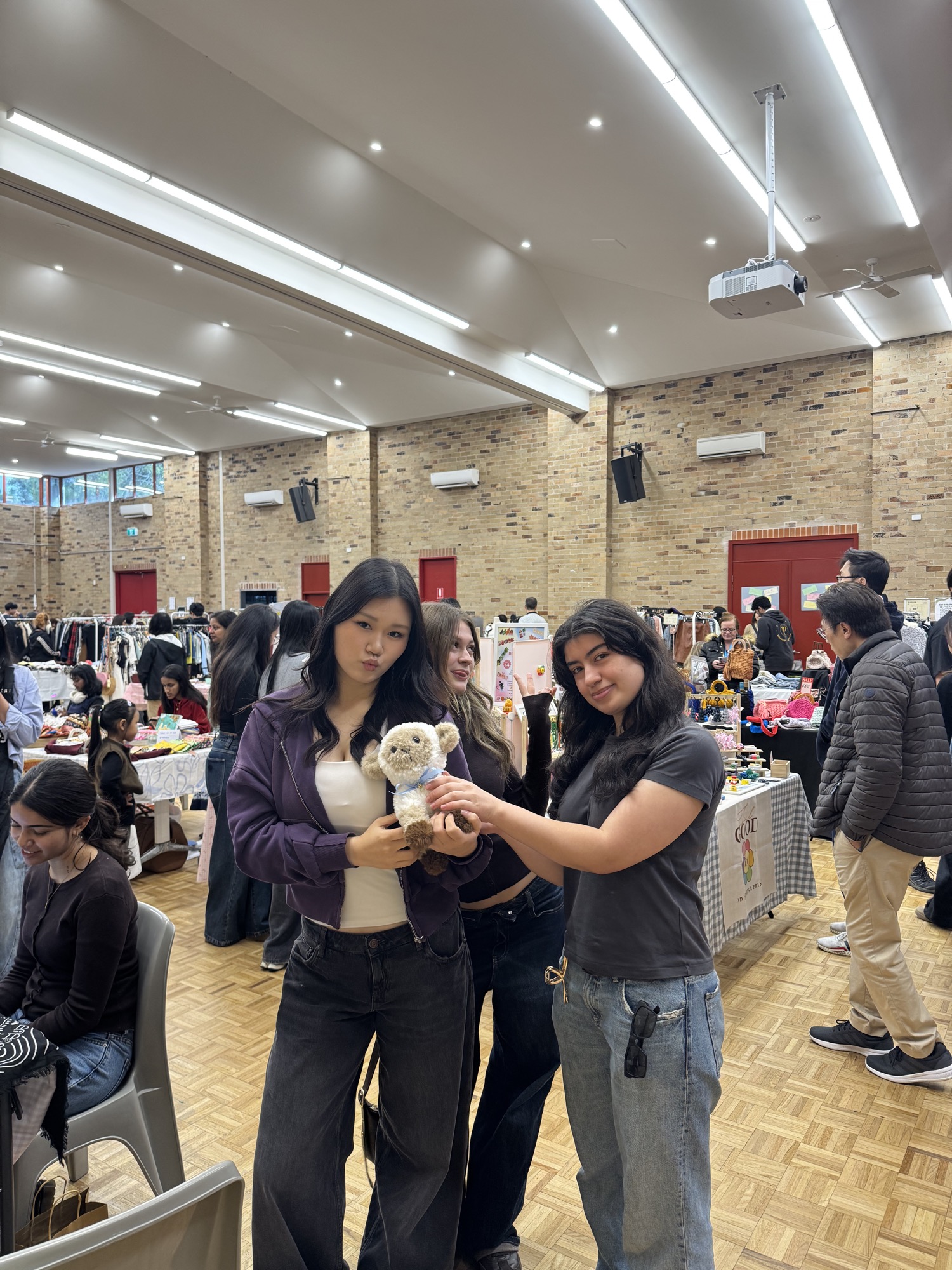 Three friends laughing together while holding a handmade teddy bear at the market