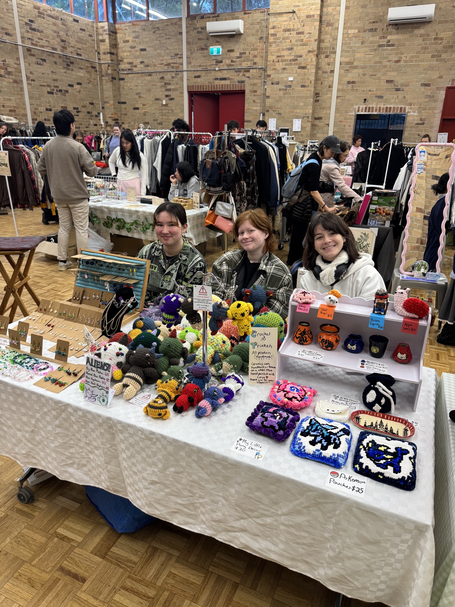 Three stallholders behind a table piled with crochet toys, trinkets and handmade ceramics