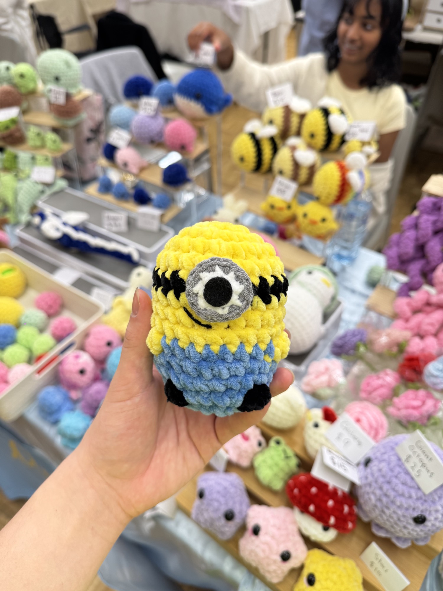 A hand holding a yellow crocheted character in front of a stall of crocheted bees, frogs, mushrooms and other toys