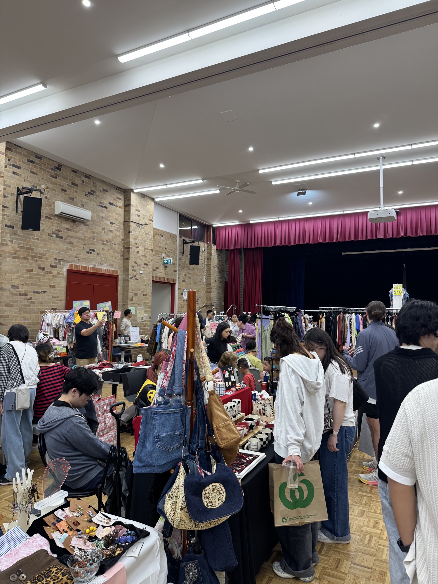 Shoppers browsing bags and jewellery beside clothing racks, with the stage curtains behind them