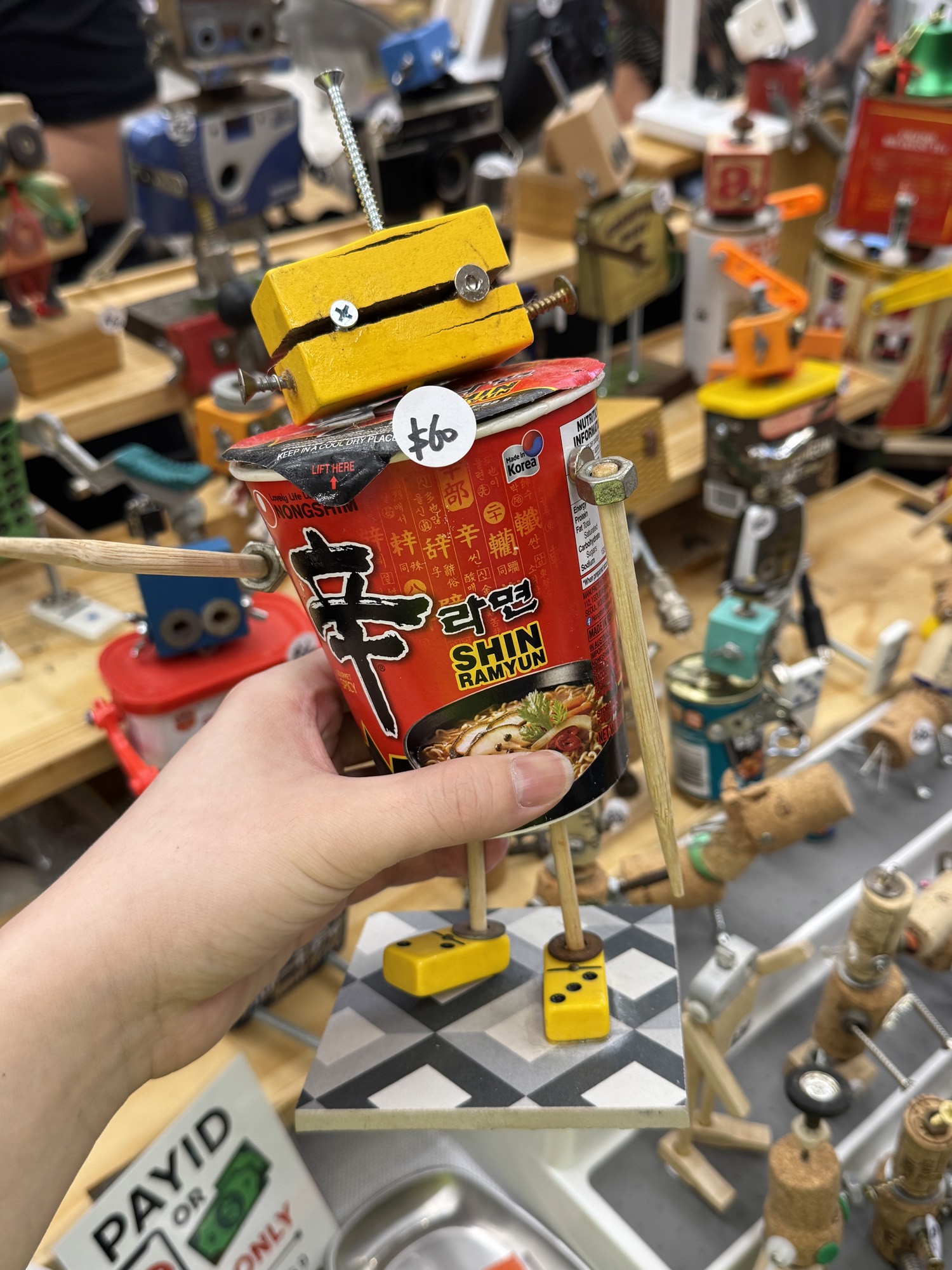 A hand holding a sculpture made from a noodle cup, chopsticks and screws, with more recycled figures on the table behind