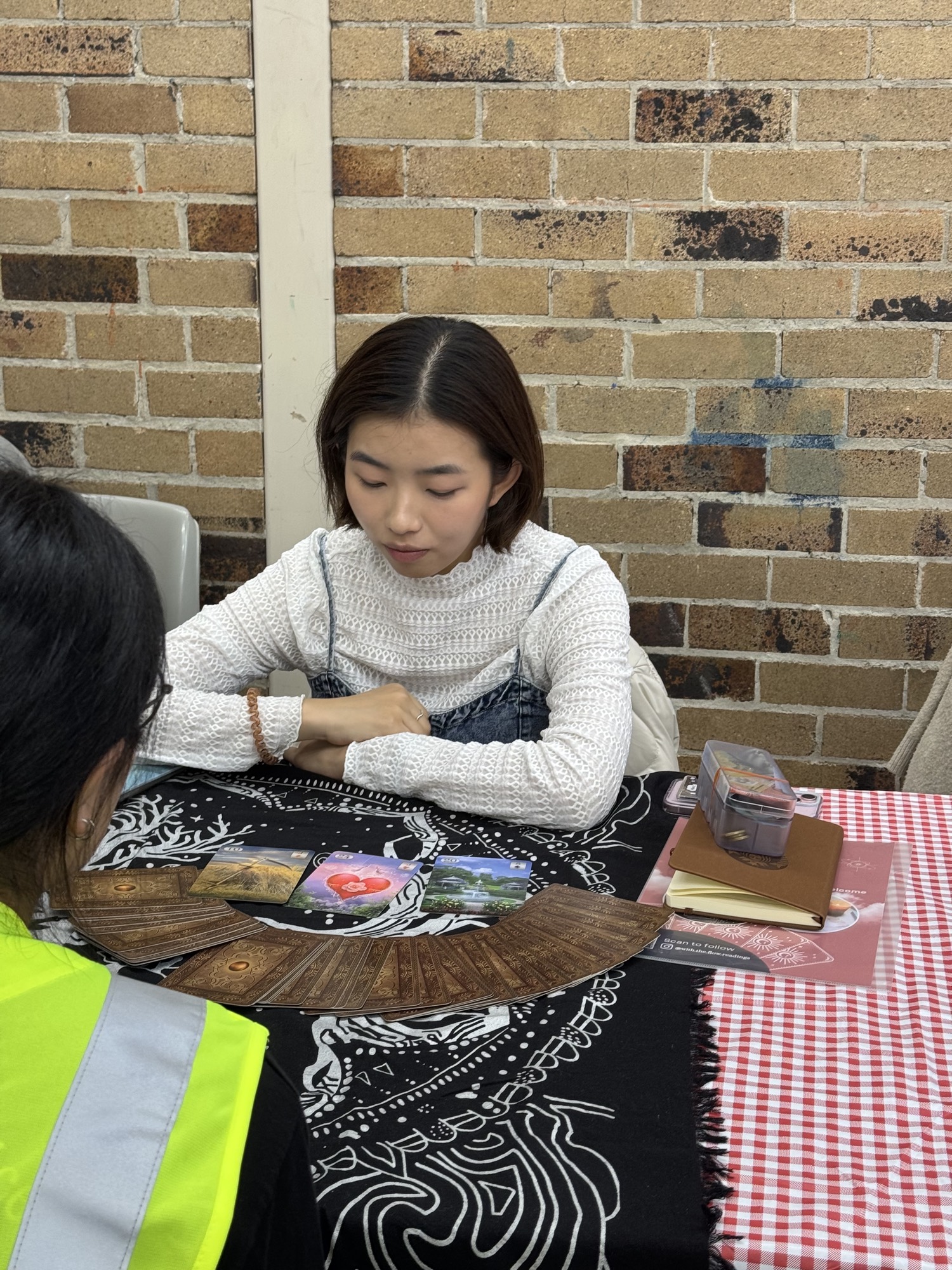 A tarot reader in a white knit top studying cards spread on a black-and-white cloth against a brick wall